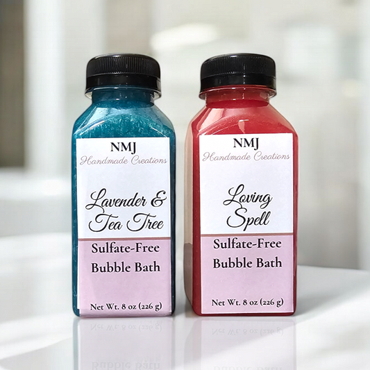 Two bottles of NMJ Handmade Creations sulfate-free bubble bath in 'Lavender & Tea Tree' and 'Loving Spell'. The first bottle is colored vibrant blue, the second rich red, both displayed against a clean, bright bathroom backdrop.