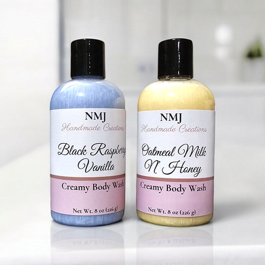 Two bottles of NMJ Handmade Creations' Creamy Body Wash, one with "Black Raspberry Vanilla" and the other "Oatmeal Milk N' Honey," displayed on a clean, white countertop against a bathroom backdrop