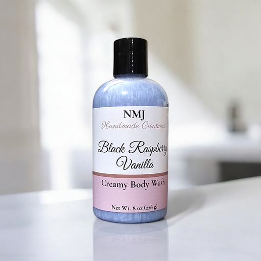 A bottle of NMJ Handmade Creations' "Black Raspberry Vanilla" Creamy Body Wash stands on a pristine, white bathroom counter. The bottle's soothing lavender color gradient adds a touch of elegance