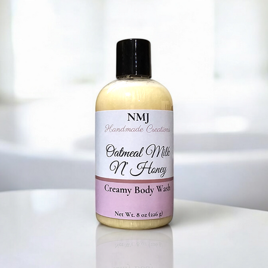 A bottle of NMJ Handmade Creations' "Oatmeal Milk N' Honey" Creamy Body Wash is displayed on a clean, white bathroom counter. The creamy, pale yellow wash visible through the clear bottle promises a soothing and nourishing experience.