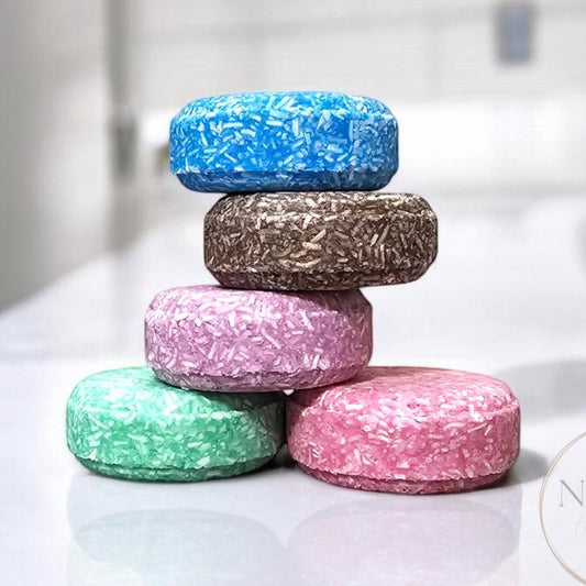 appealing stack of multicolored conditioner bars on a glossy white surface. The bars are in shades of blue, brown, pink, green, and lighter pink, each with a unique texture resembling tiny strands, giving a vibrant and eco-friendly aesthetic.