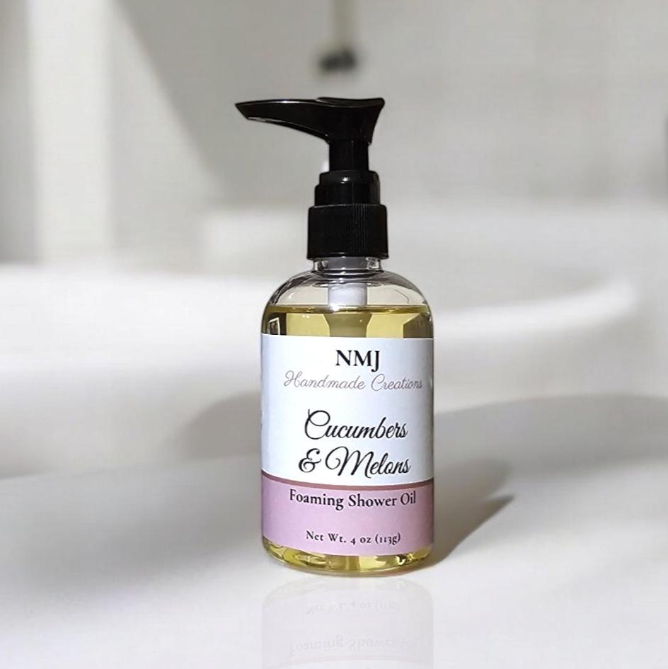 A bottle of NMJ Handmade Creations' "Cucumbers & Melons" foaming shower oil is displayed against a minimalist bathroom backdrop. The label features a refreshing design with soft pink and white tones, emphasizing the crisp and invigorating scent of the product