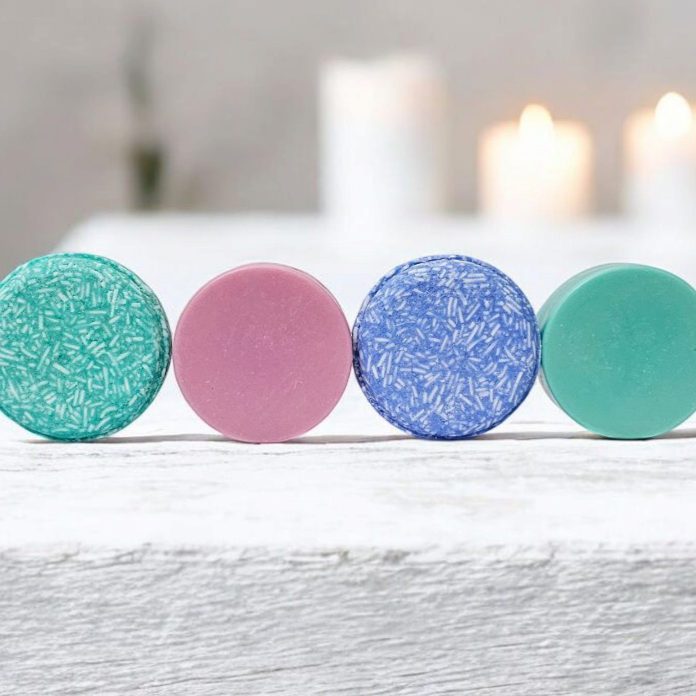 Four vibrant shampoo bars in a line on a distressed white wooden surface, with lit candles softly glowing in the background. From left to right, the bars are teal with textured shreds, solid matte pink, textured blue, and smooth aqua. The arrangement showcases the variety of colors and textures, highlighting the natural and artisanal quality of each bar